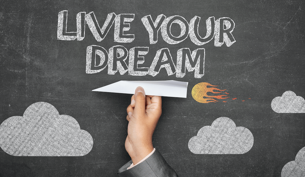 Hand holding a paper airplane in fromt of a chalkboard that has Live Your Dream written on it